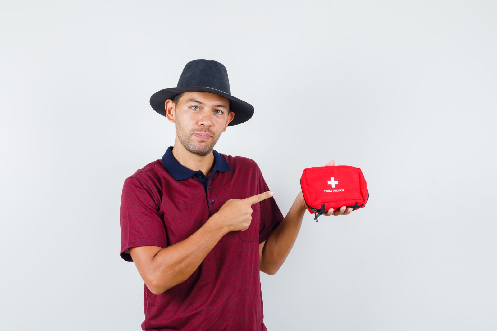 Young male pointing to fast aid kit in red shirt,black hat and looking serious , front view.